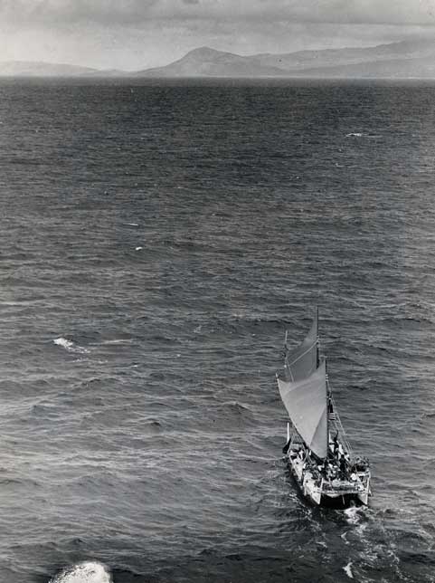 Hokulea in the foreground on the ocean. Photo is black and white. On the ocean's horizon you see the shadow of an island.